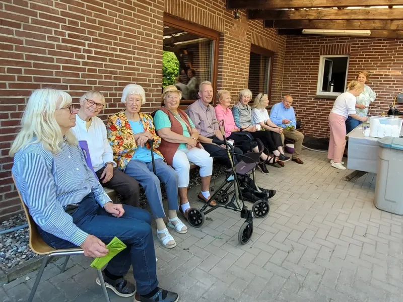 Ältere Menschen sitzen bei einem geselligen Beisammensein auf einer Terrasse unter einer Holzpergola.
