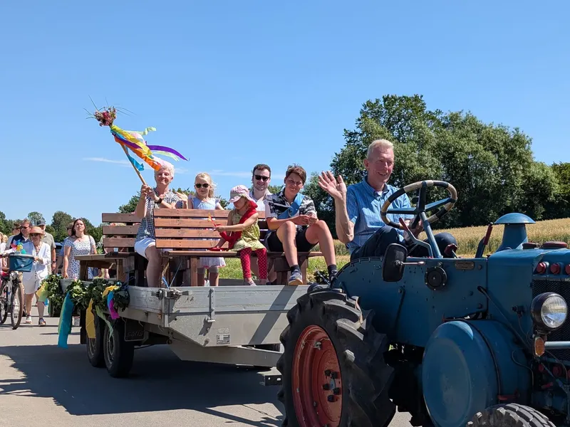 Ein Mann fährt bei einem Festumzug einen blauen Oldtimer-Traktor mit einem geschmückten Anhänger voller feiernder Menschen.